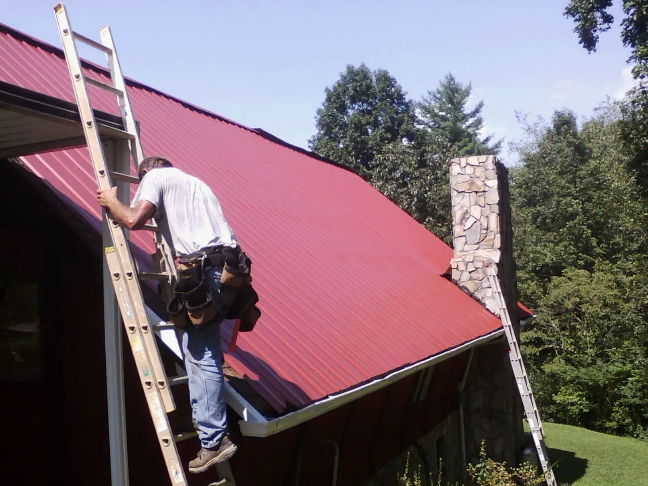 Roofer on a ladder beside a red metal roof and stone chimney — Soffit & Fascia Repair by Roofing Brothers in Trout Creek
